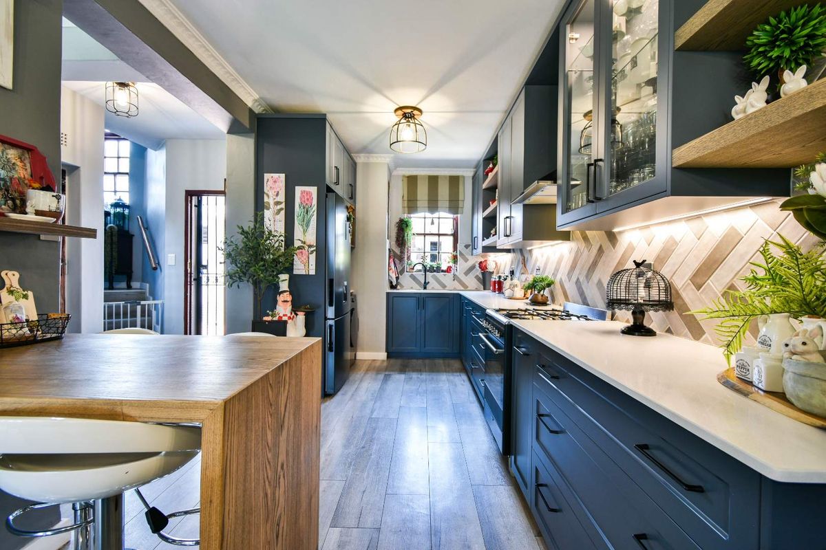 Modern galley-style kitchen featuring dark cabinetry, stone countertops, herringbone tile backsplash, and integrated appliances, leading into an adjacent living area.
