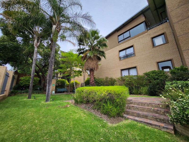 A well-maintained rear garden area featuring a brick apartment facade, mature palm trees, landscaped hedges, and brick steps leading to the building.