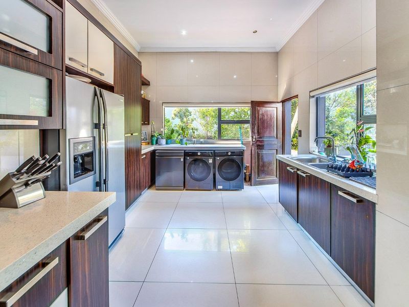 A modern, well-lit scullery featuring dark wood cabinetry, stone countertops, a double sink, and large windows overlooking the garden.