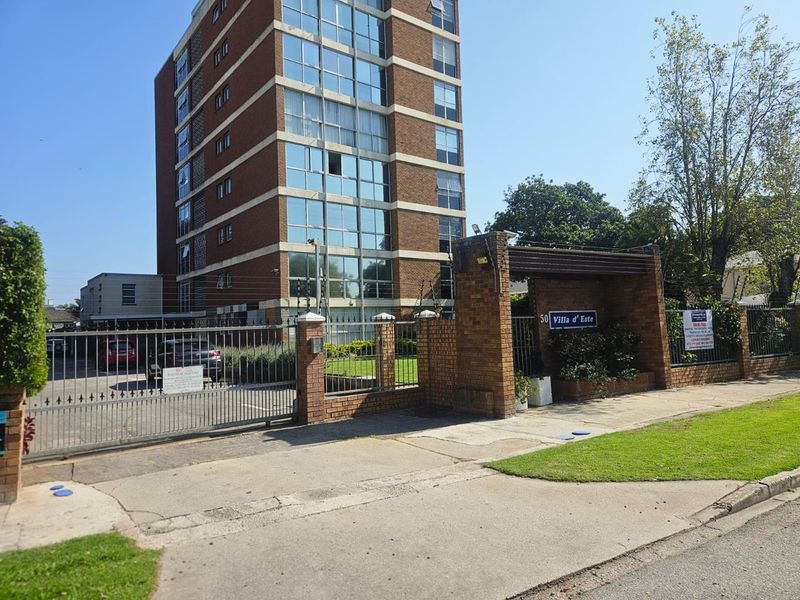 Front exterior view of Villa d'Este apartment complex featuring a brick perimeter wall, metal security gate, and multi-story building with glass facade.