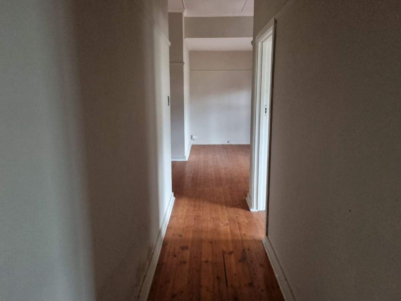 A vacant hallway featuring timber flooring and neutral walls, leading into an adjacent room. The space shows signs of wear and requires cosmetic attention.