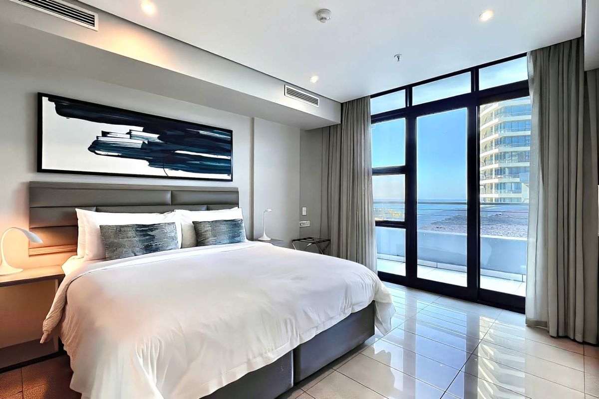 Modern bedroom featuring tiled flooring, recessed ceiling lighting, ducted air conditioning, and large glass sliding doors opening to a private balcony with ocean views.