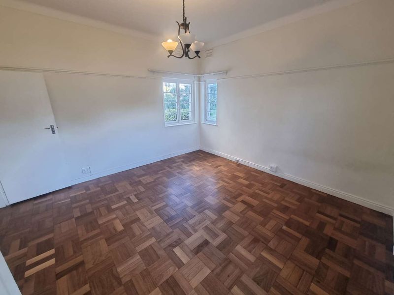 A bright, empty bedroom featuring classic parquet flooring, picture rails, and corner windows with security bars.