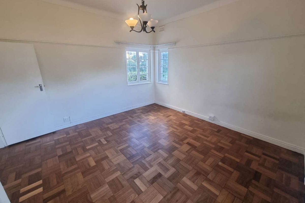 A bright, empty bedroom featuring classic parquet flooring, picture rails, and corner windows with security bars.