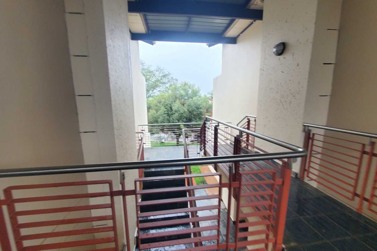 An exterior view of an apartment complex walkway featuring metal railings, tiled flooring, and a covered overhead structure overlooking the surrounding greenery.