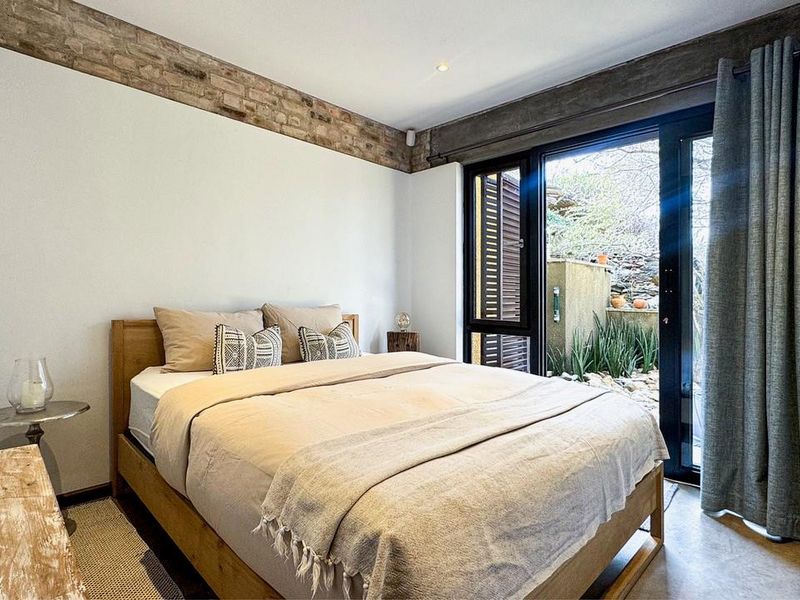 A modern bedroom featuring exposed brick accents, a concrete ceiling beam, polished concrete floors, and direct access to a private outdoor garden area.