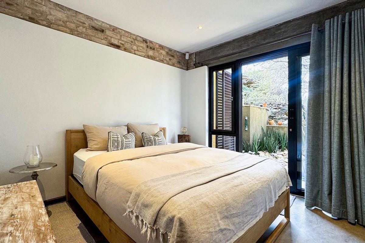 A modern bedroom featuring exposed brick accents, a concrete ceiling beam, polished concrete floors, and direct access to a private outdoor garden area.
