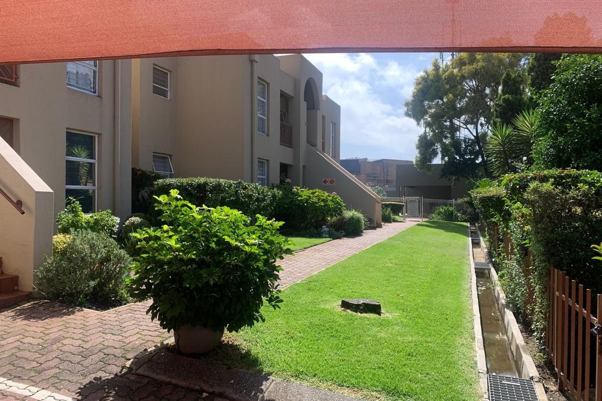 A well-maintained communal garden area featuring a paved walkway, manicured lawn, and established landscaping, viewed from a shaded patio area.