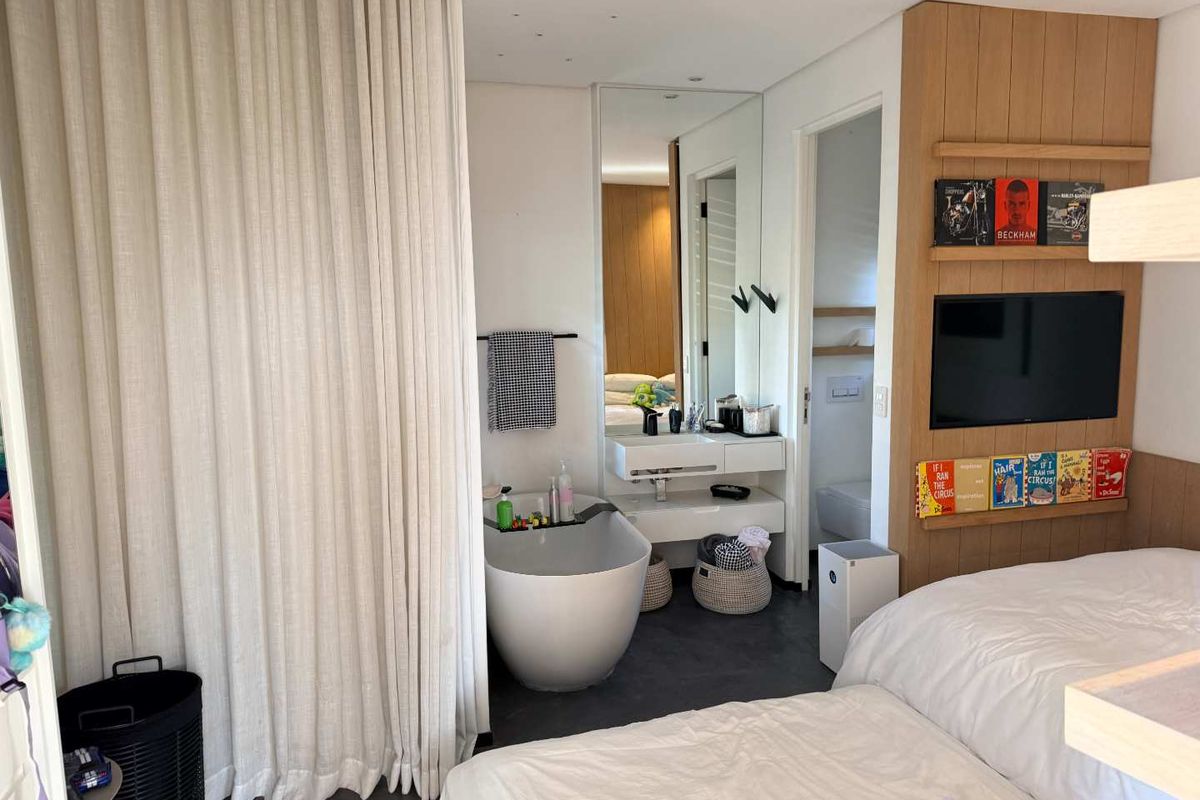 Modern bedroom featuring an open-plan ensuite with a sleek freestanding bathtub, wall-mounted vanity, and integrated timber shelving with a built-in television.