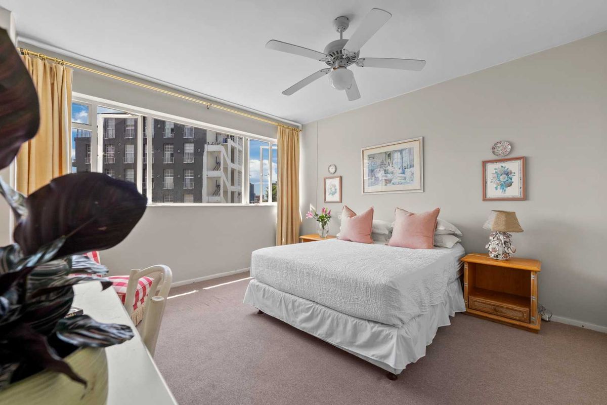 A well-lit bedroom featuring neutral walls, carpeted flooring, a ceiling fan, and a large window providing natural light and urban views.