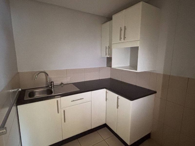 A clean and functional kitchenette featuring white built-in cabinetry, dark laminate countertops, a stainless steel sink, and neutral tiled splashbacks.