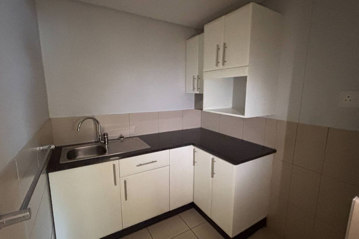 A clean and functional kitchenette featuring white built-in cabinetry, dark laminate countertops, a stainless steel sink, and neutral tiled splashbacks.