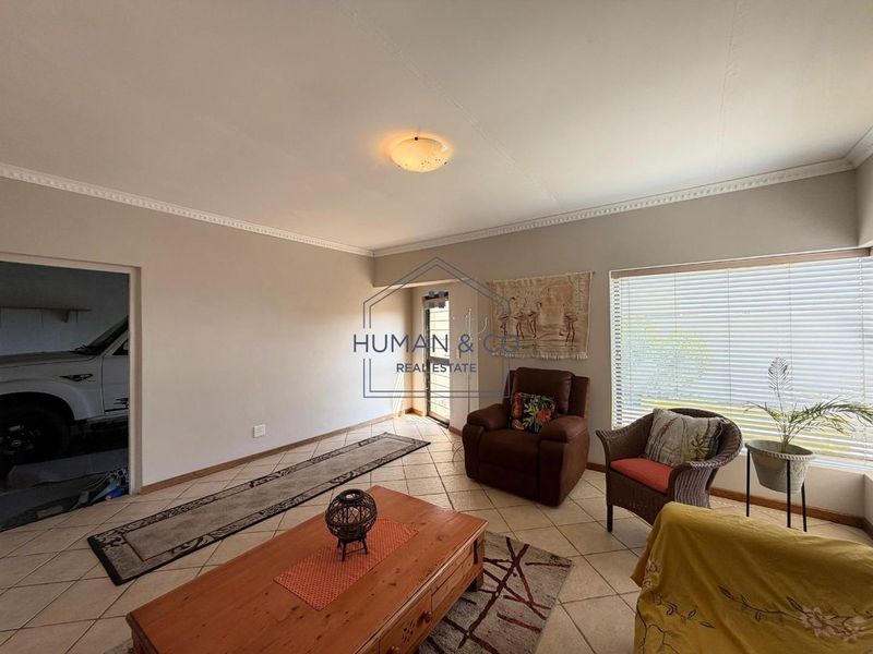 A spacious living room featuring neutral wall tones, tiled flooring, decorative ceiling cornices, and large windows fitted with venetian blinds.