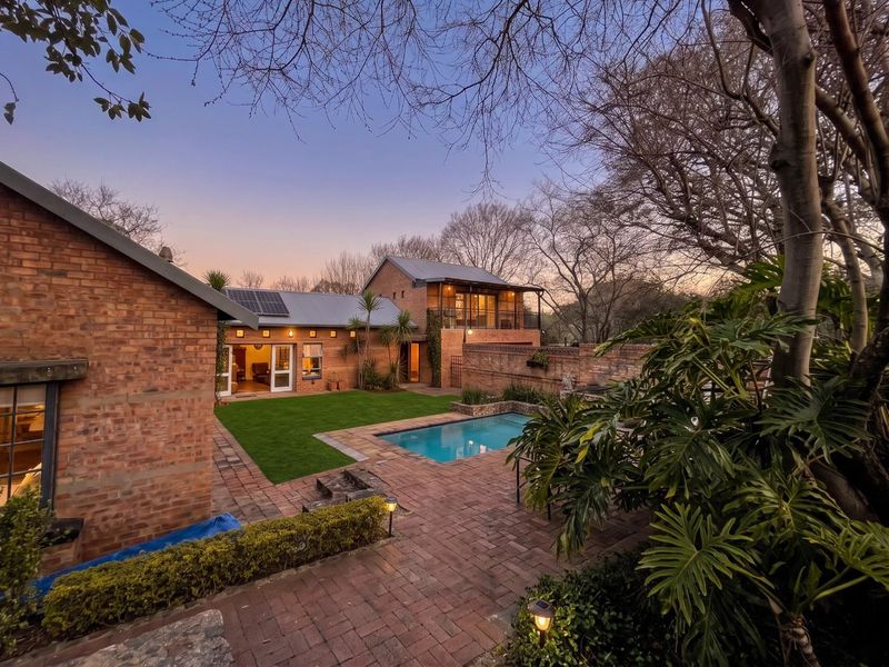 A beautiful rear exterior view featuring a swimming pool, brick-paved patio, lush lawn, and a multi-level brick house with a balcony at dusk.