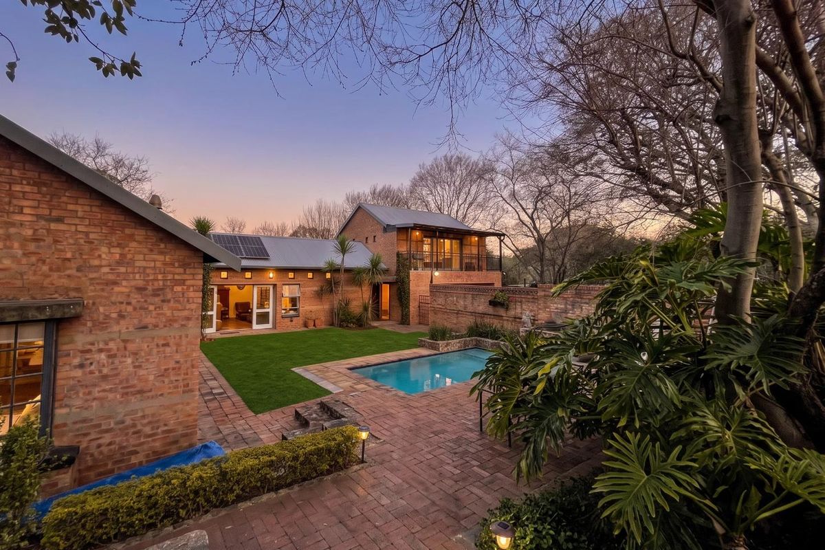 A beautiful rear exterior view featuring a swimming pool, brick-paved patio, lush lawn, and a multi-level brick house with a balcony at dusk.