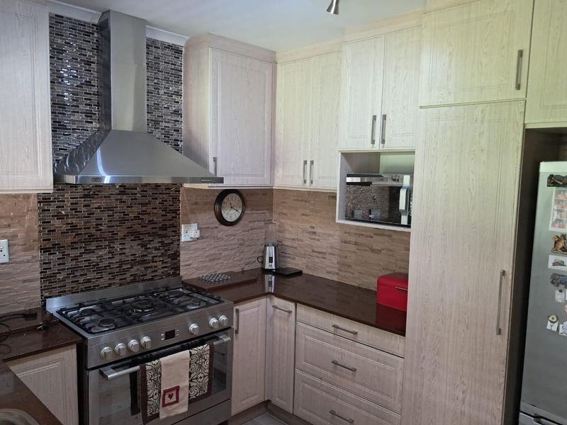 A well-maintained kitchen featuring light wood-grain cabinetry, stone countertops, a gas stove with stainless steel extractor fan, and a decorative mosaic tile backsplash.