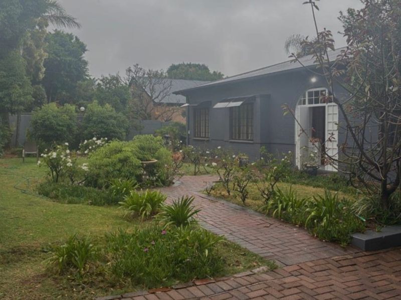 A view of the rear garden featuring a brick paved pathway leading to the house entrance, surrounded by established landscaping and lawn.