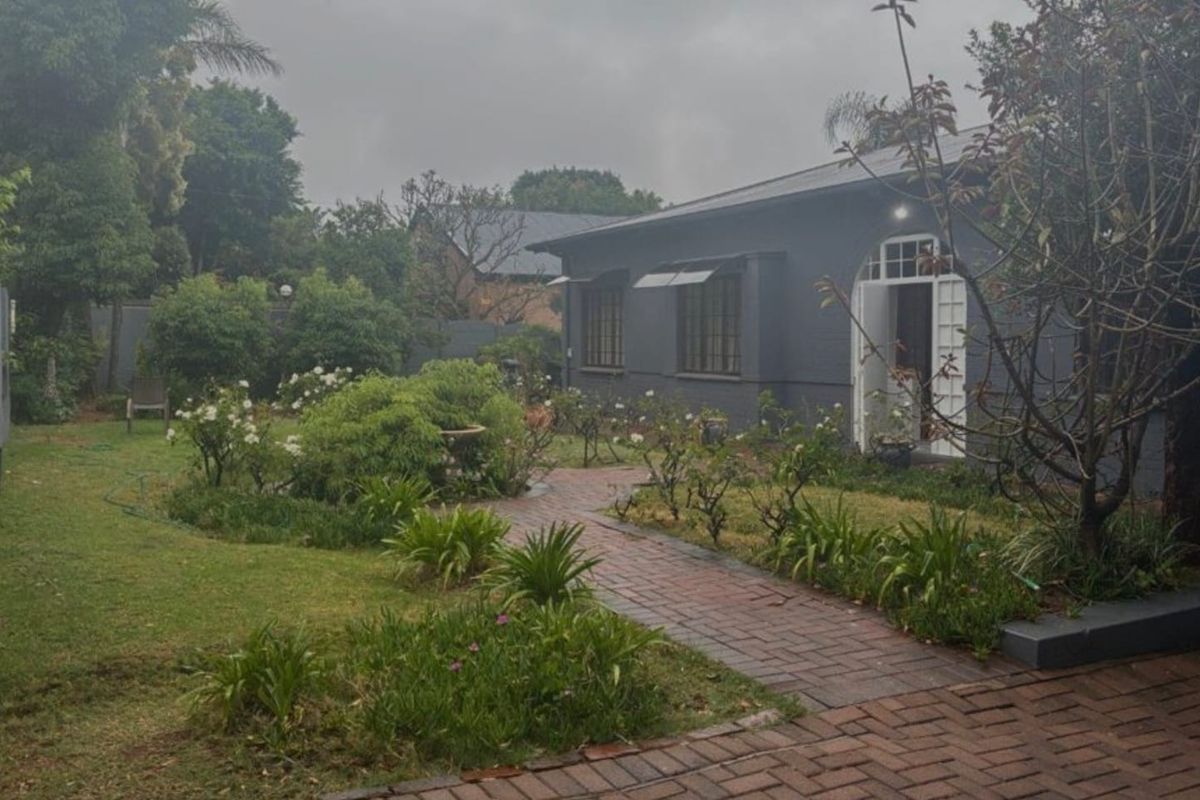 A view of the rear garden featuring a brick paved pathway leading to the house entrance, surrounded by established landscaping and lawn.