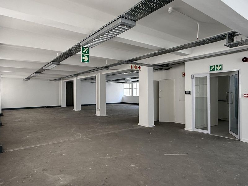 Spacious open-plan office area featuring exposed industrial ceilings, concrete flooring, and integrated emergency safety systems, offering a versatile blank canvas for business operations.