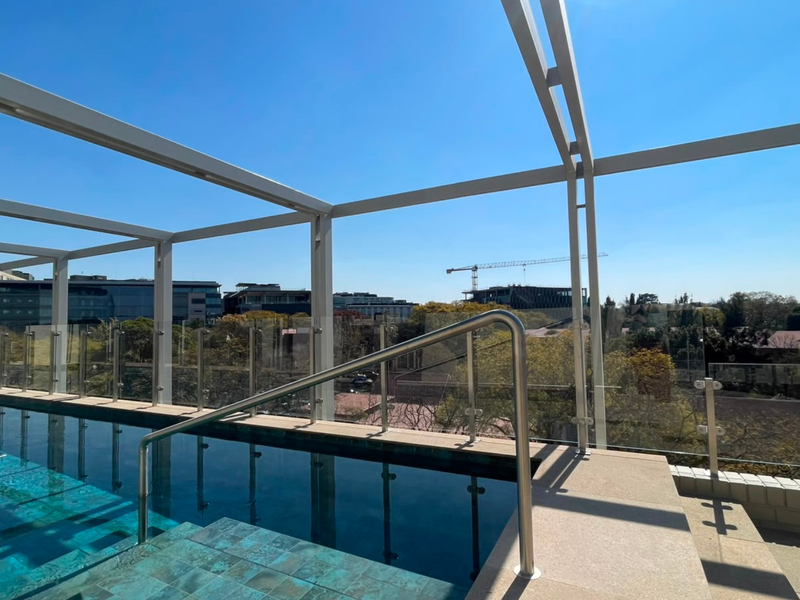 A modern rooftop swimming pool featuring a glass balustrade, metal pergola structure, and stainless steel handrail, offering clear views of the surrounding urban landscape.