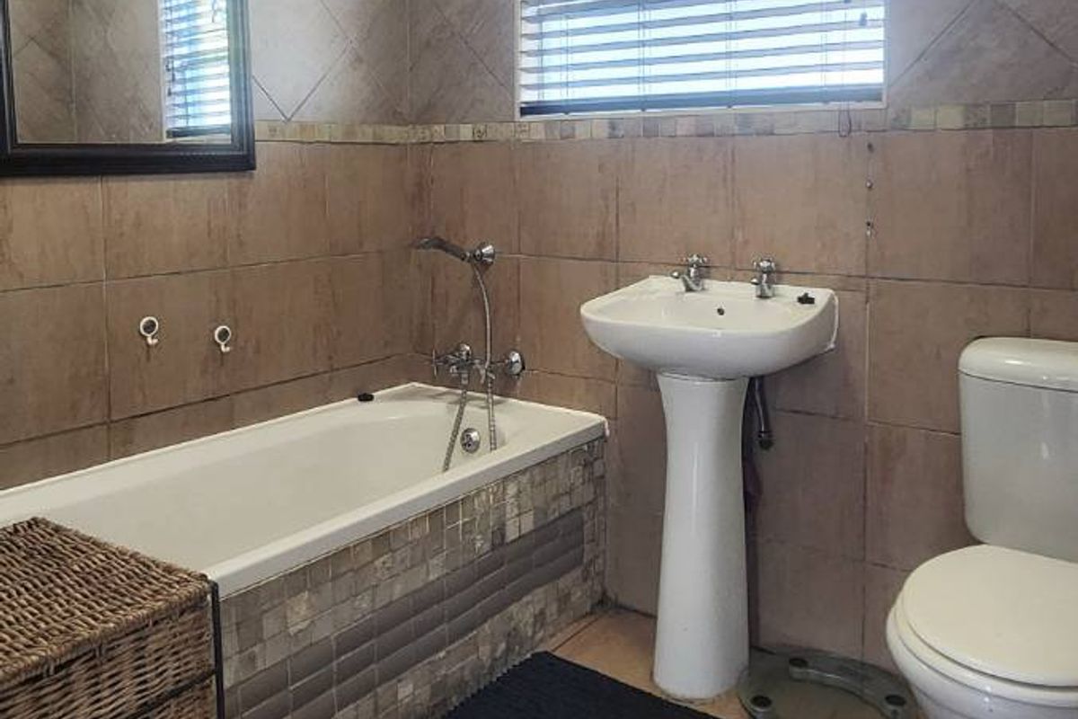 A functional bathroom featuring a bathtub, pedestal basin, and toilet, finished with neutral wall and floor tiling and a window with venetian blinds.