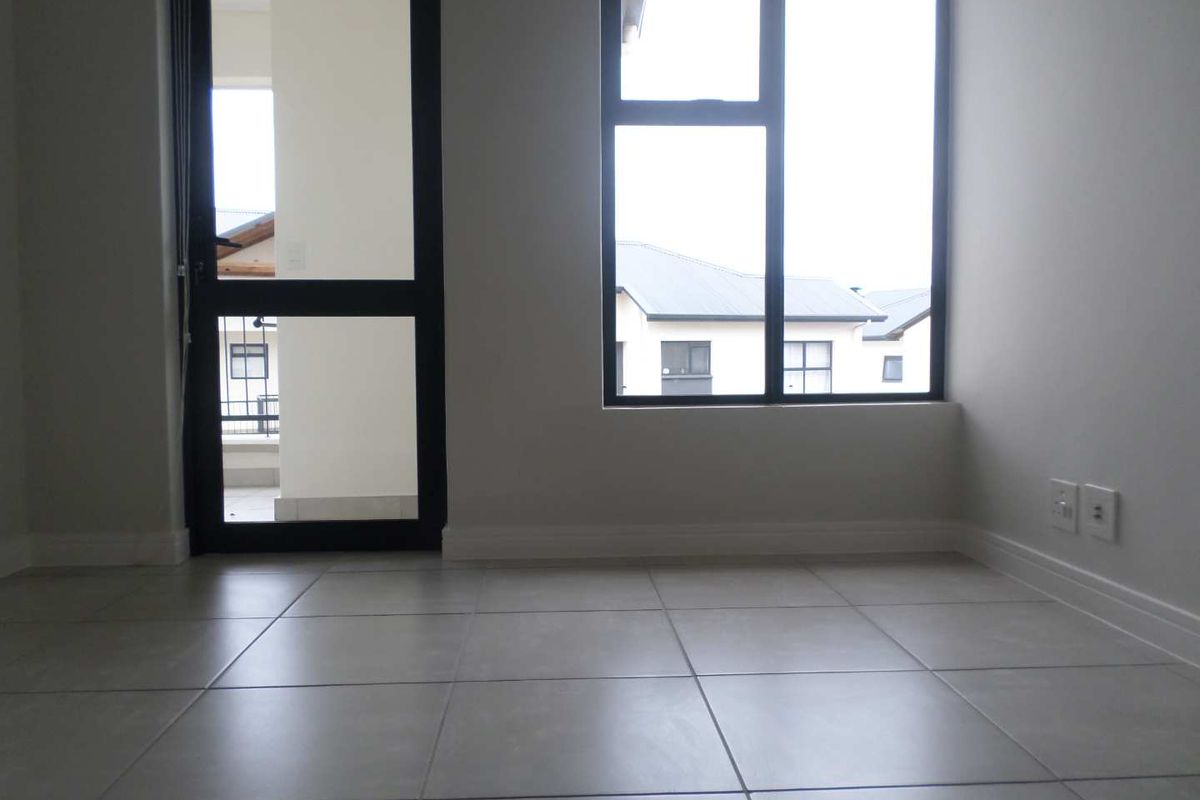 A bright, empty room featuring neutral floor tiles, large windows with dark frames, and a glass door leading to an outdoor balcony area.