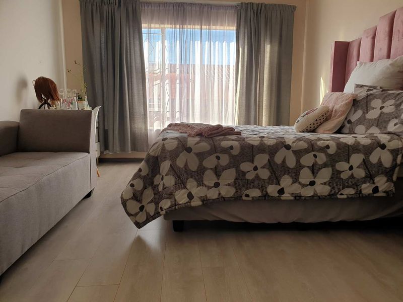 A well-lit bedroom featuring light-toned laminate flooring and large windows with curtain rails, providing a comfortable and functional living space.