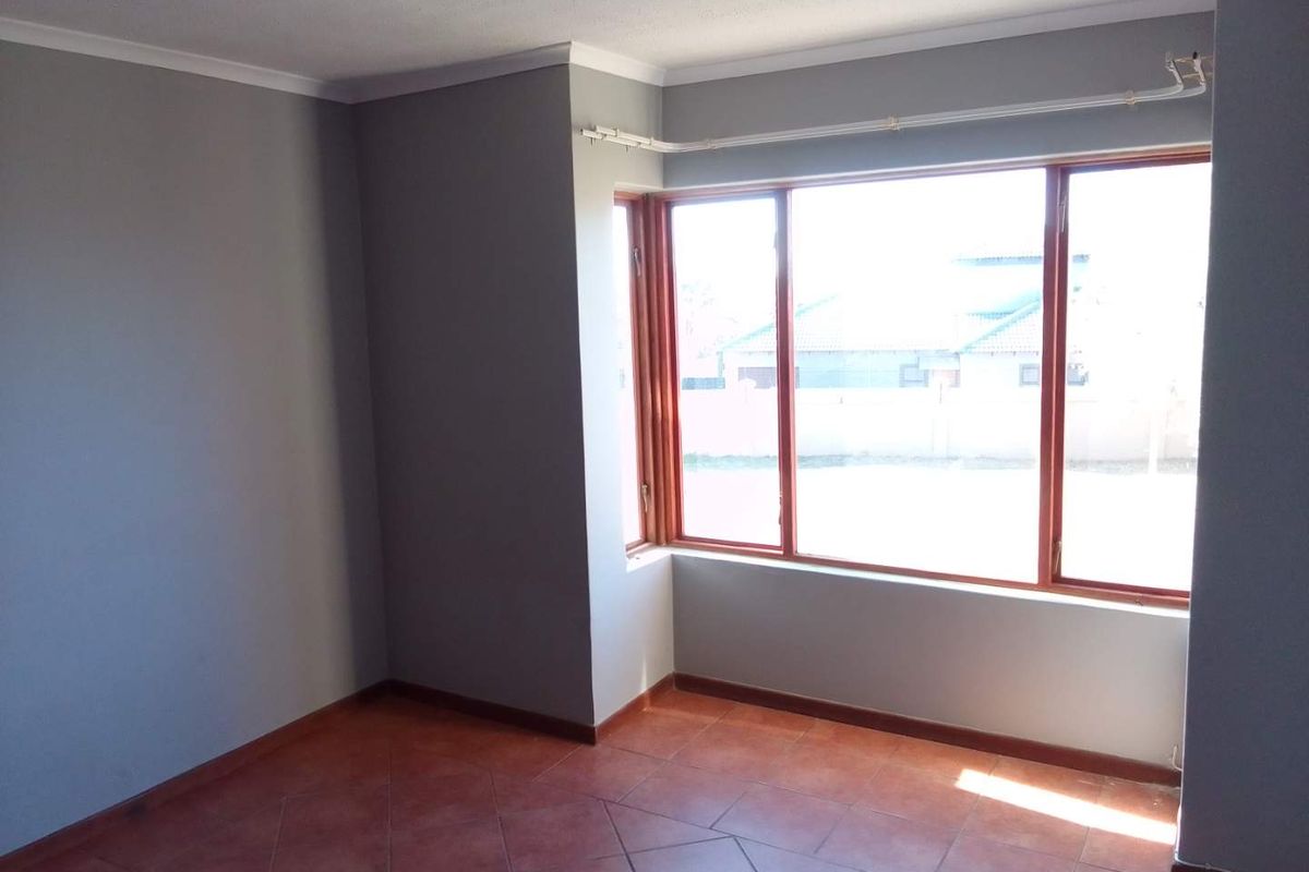 A bright, empty bedroom featuring terracotta-style floor tiles, grey painted walls, and large windows with wooden frames.