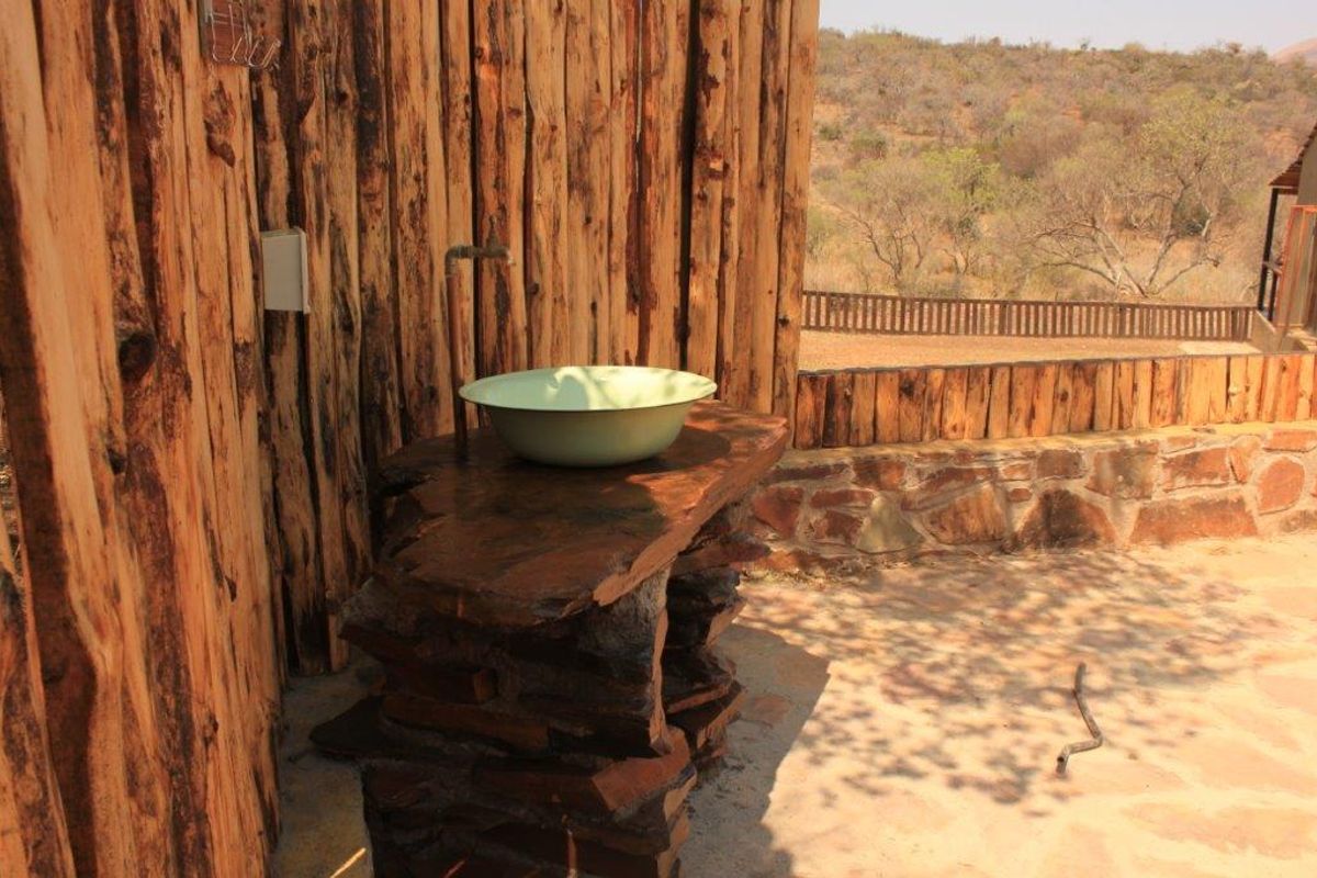 An outdoor bathroom area featuring a rustic stone vanity with a vessel sink, set against a natural timber log wall and stone paving.