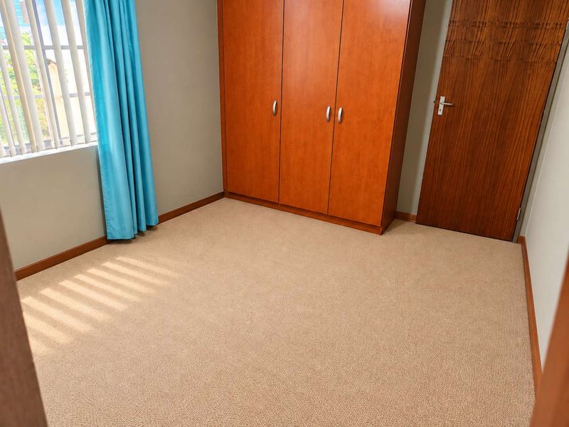 A clean, empty bedroom featuring neutral carpet flooring, a built-in wooden wardrobe, and a window with venetian blinds.