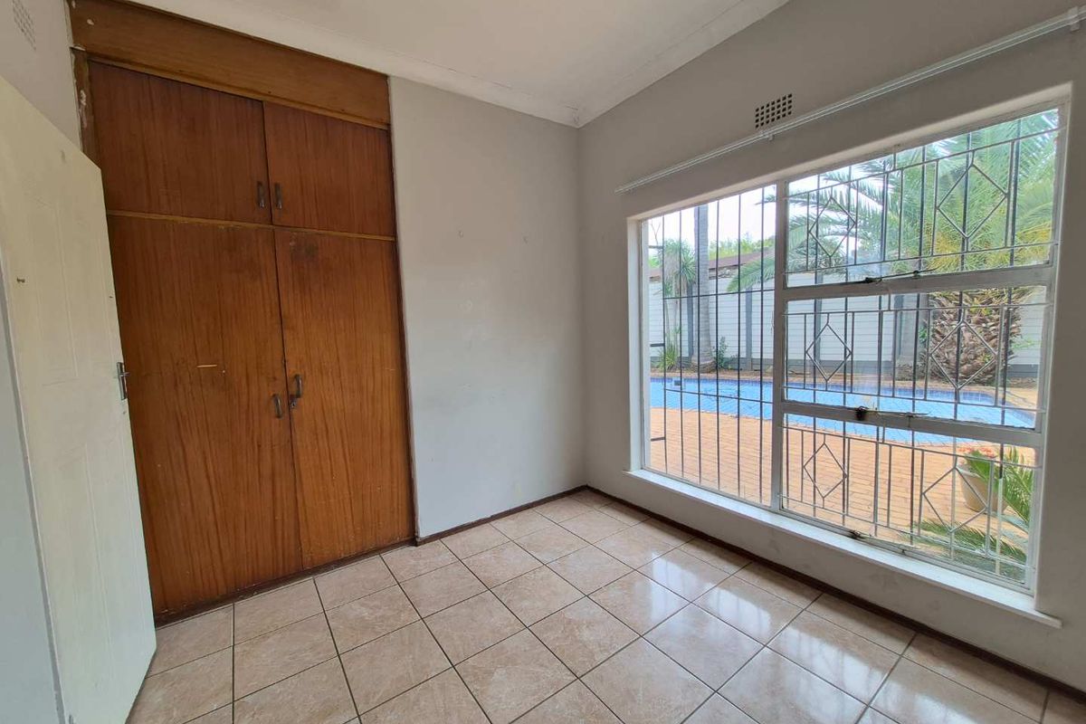 A vacant bedroom featuring tiled flooring, built-in wooden cupboards, and a large window with security bars overlooking the pool area.