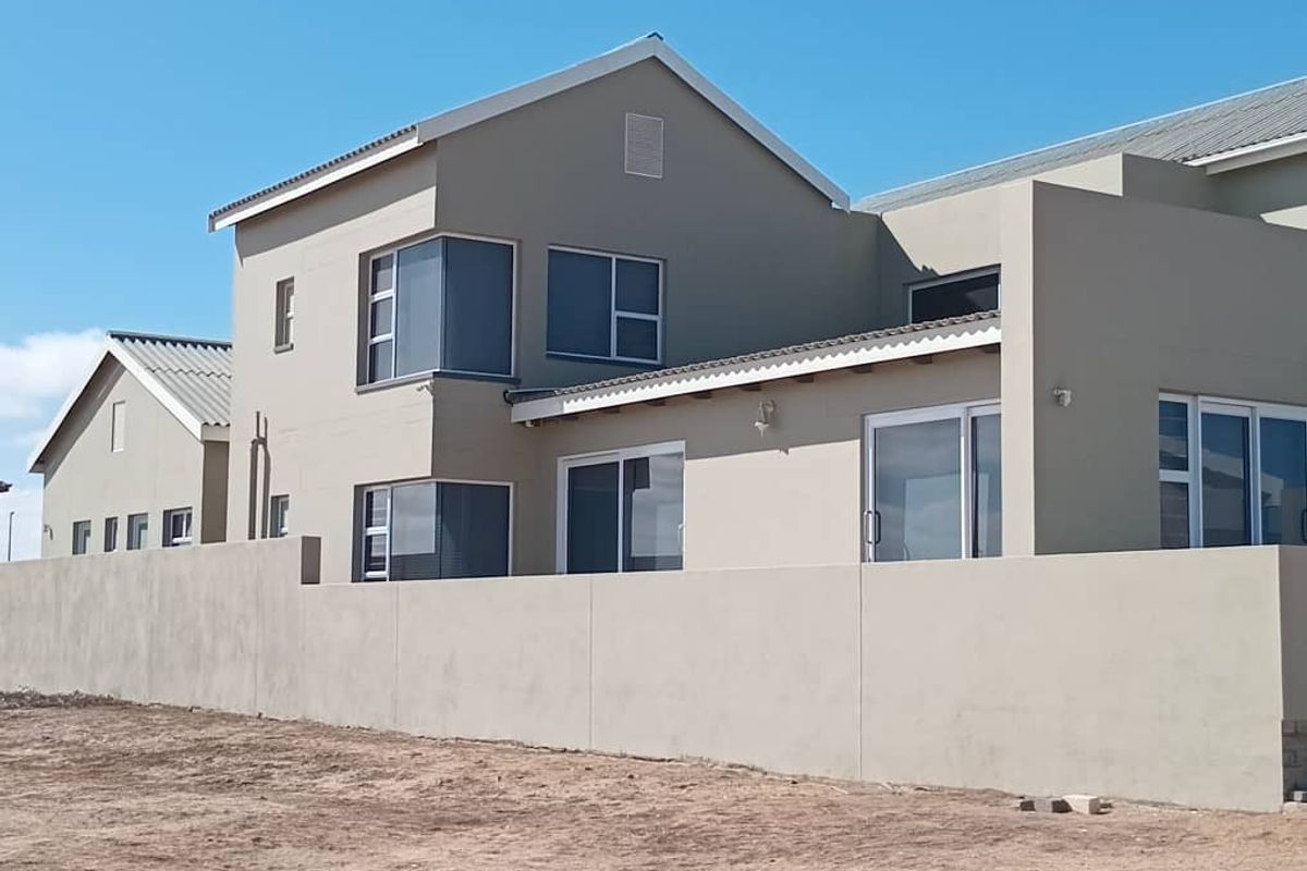 A view of the rear exterior of a modern double-story house featuring neutral-toned plaster walls, sliding glass doors, and a perimeter boundary wall.