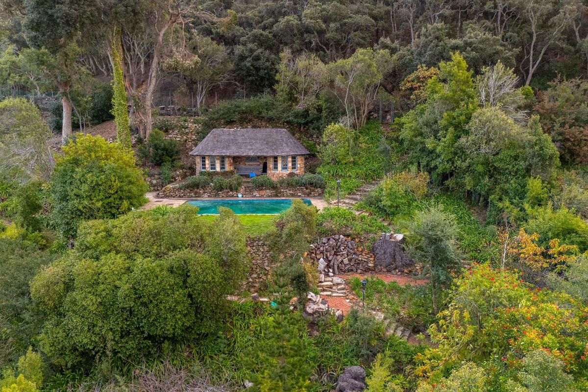 An elevated aerial view of a secluded residential property featuring a stone cottage, a swimming pool, and lush, mature garden landscaping.