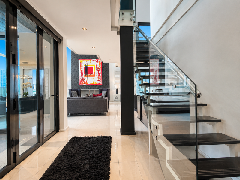 Modern hallway featuring a sleek glass balustrade staircase, polished tiled flooring, and large sliding glass doors leading to an outdoor area.