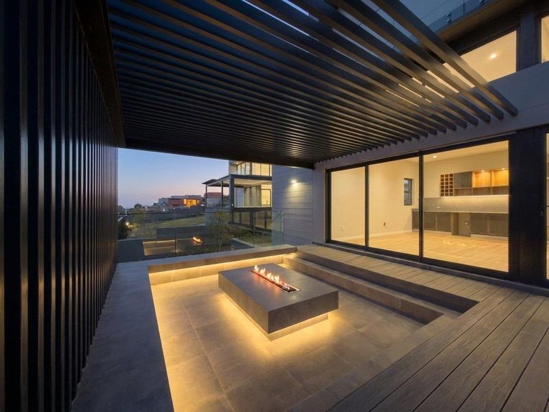 Modern covered patio featuring a built-in fire pit, slatted pergola, glass balustrade, and seamless indoor-outdoor flow through large sliding glass doors.