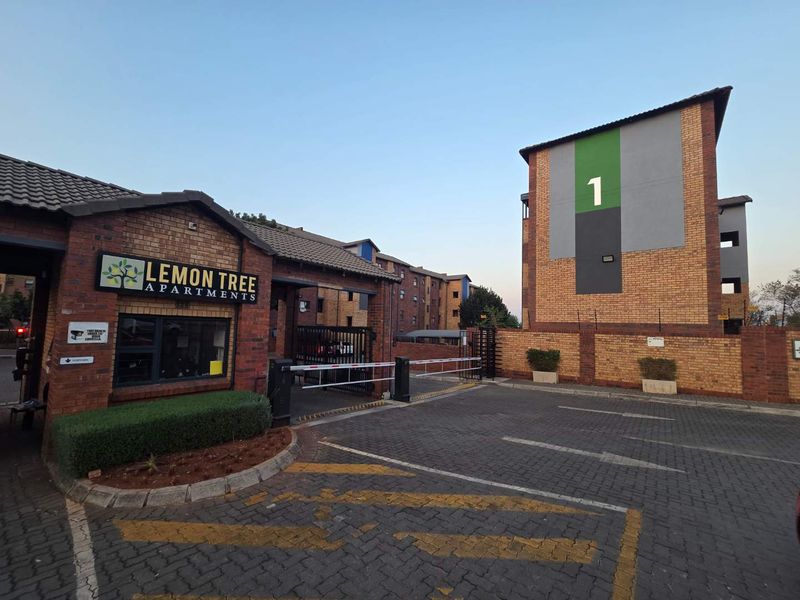 The main entrance to Lemon Tree Apartments featuring a secure boom gate, brick facade buildings, and a paved driveway.