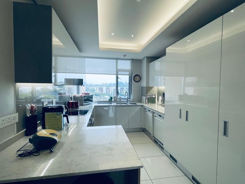 Modern kitchen featuring sleek white cabinetry, stone countertops, integrated appliances, and recessed ceiling lighting with a large window providing natural light and views.