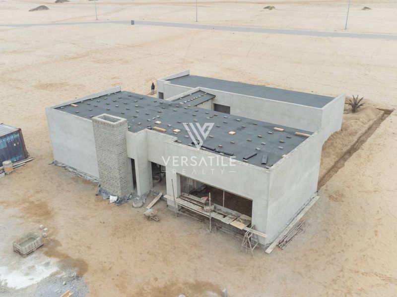 Aerial view of a residential property currently under construction, featuring a concrete shell and flat roof design in a sandy landscape.