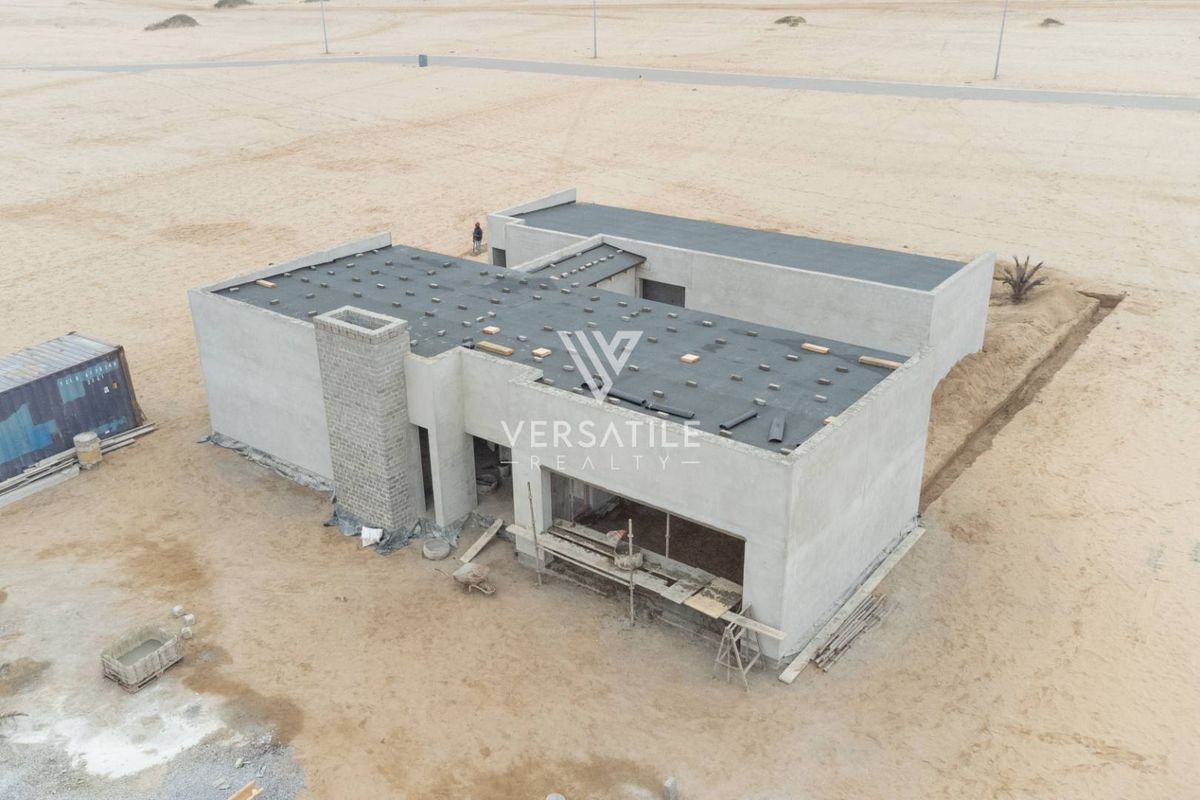 Aerial view of a residential property currently under construction, featuring a concrete shell and flat roof design in a sandy landscape.