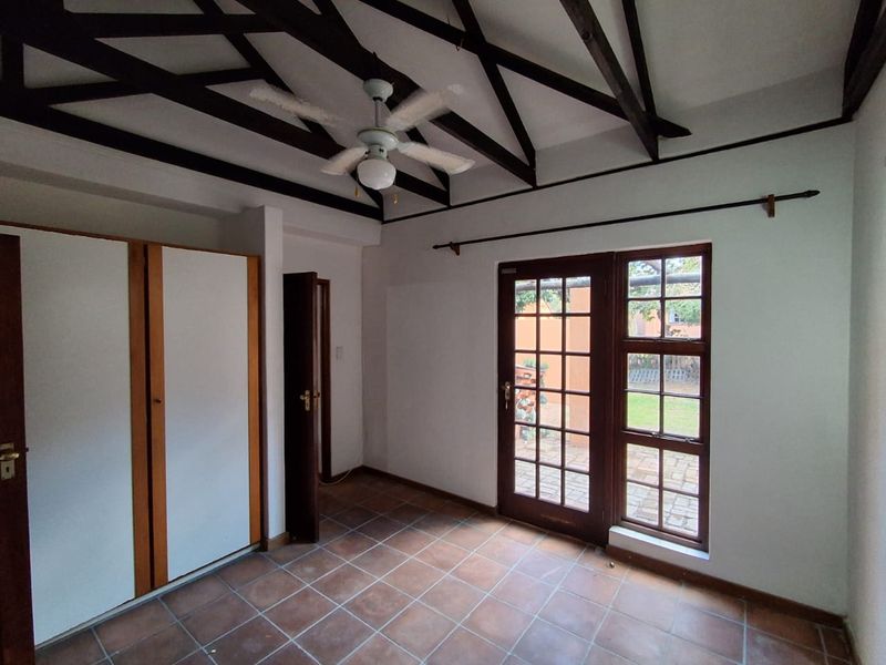 A bedroom featuring exposed timber ceiling beams, built-in cupboards, tiled flooring, and direct access to the garden through a timber-framed glass door.
