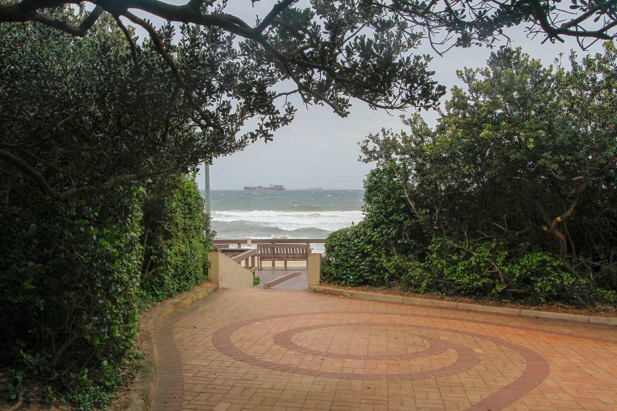 A paved outdoor walkway leading towards a coastal view, framed by mature trees and shrubs, offering direct access to the beach area.