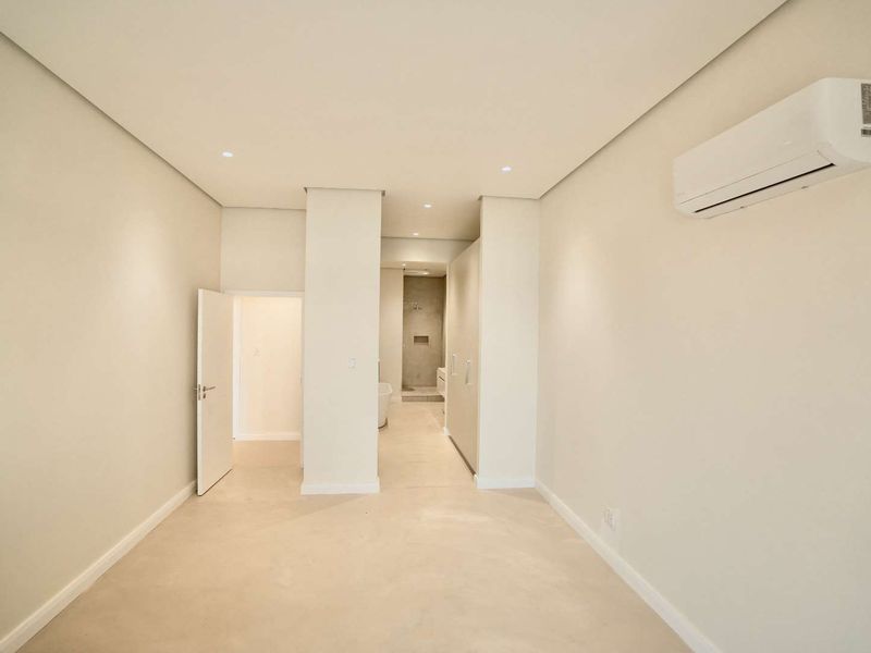 A bright, minimalist bedroom featuring smooth flooring, recessed lighting, an integrated air conditioning unit, and direct access to an en-suite bathroom.