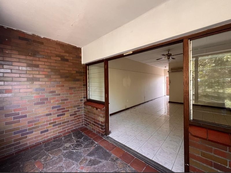 A covered patio area featuring exposed brick walls and stone flooring, leading into an open-plan interior space with tiled floors and a ceiling fan.