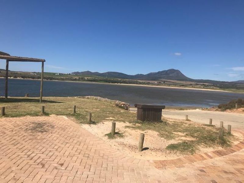 A scenic view of the waterfront featuring a paved driveway, wooden pergola structure, and natural landscape overlooking the lagoon and distant mountains.