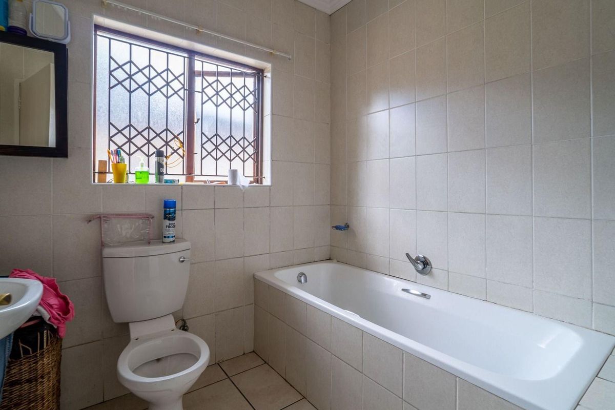 A functional bathroom featuring a built-in bathtub, toilet, and basin with neutral wall and floor tiling, complemented by a window with security bars.