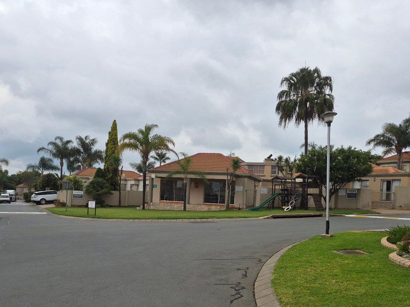 A view of the residential complex entrance featuring paved roads, landscaped gardens, and established trees under an overcast sky.