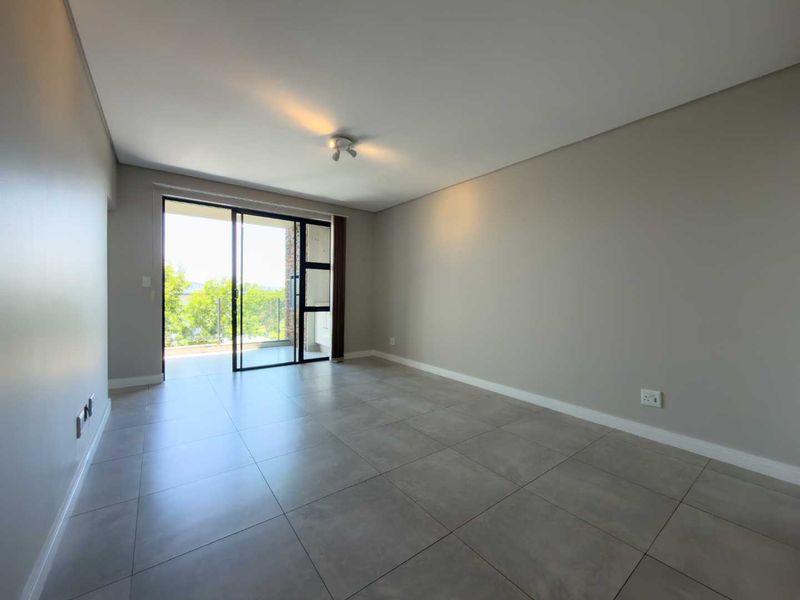 An empty, well-maintained living room featuring neutral grey floor tiles, smooth walls, and a large sliding glass door leading to an outdoor balcony.