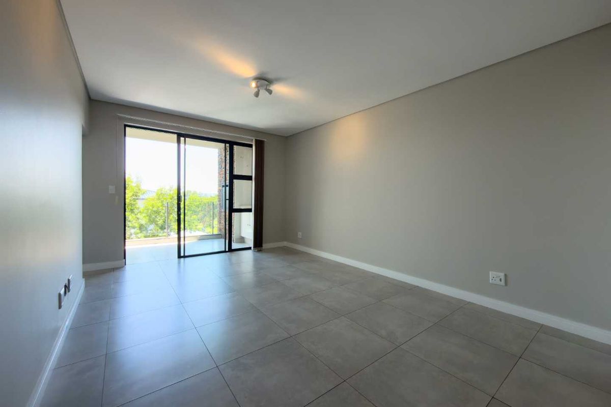 An empty, well-maintained living room featuring neutral grey floor tiles, smooth walls, and a large sliding glass door leading to an outdoor balcony.