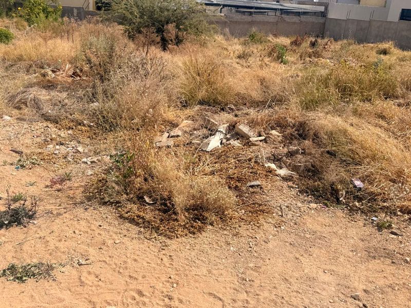 A vacant residential plot featuring dry, overgrown vegetation, scattered debris, and a perimeter boundary wall with electric fencing.