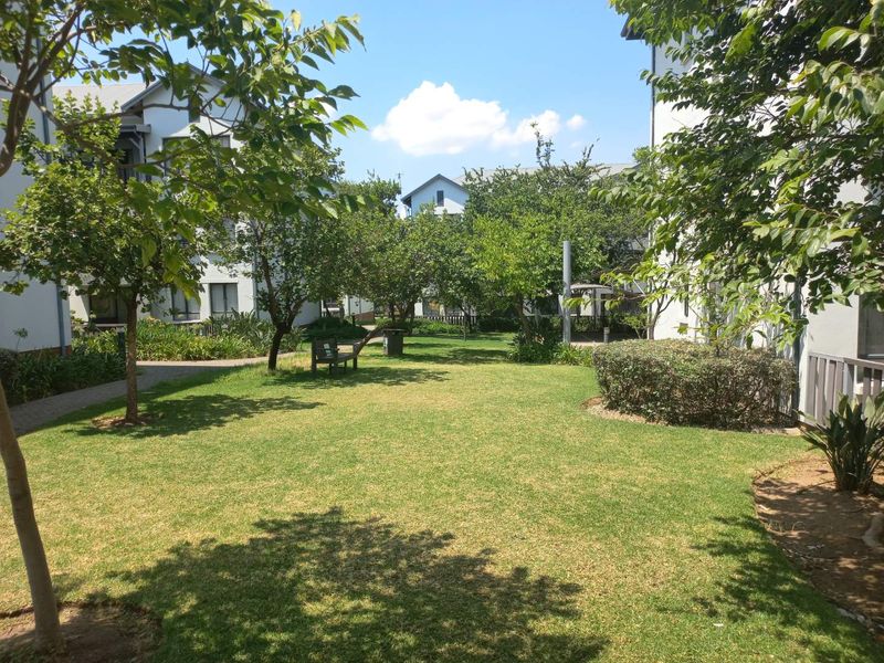 A well-maintained communal garden area featuring a manicured lawn, mature trees, and paved walkways between apartment buildings.